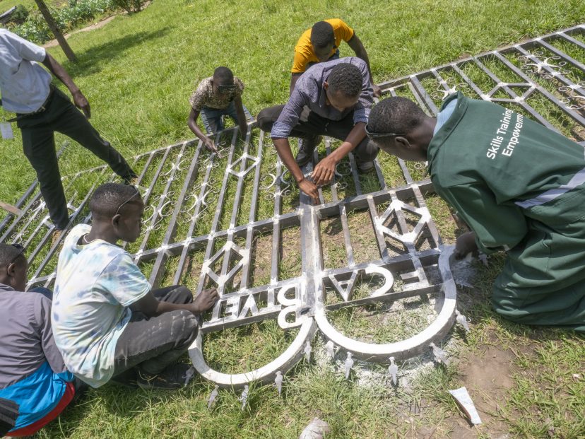 Metalwork trainees working on new main gate in Rwamwamjas. The gate is on the grass and the trainees paint it.